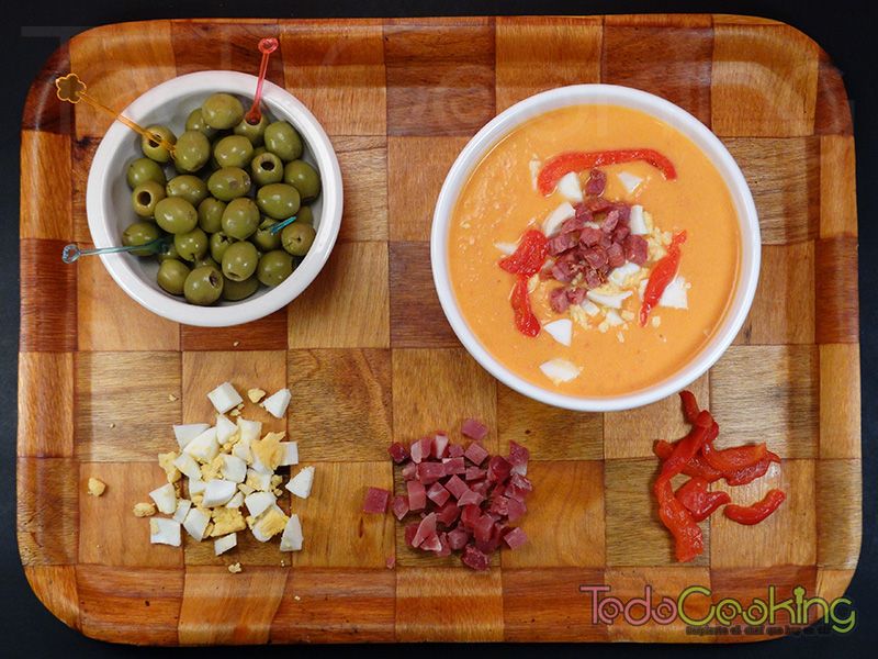 Cuenco de salmorejo cordobés rodeado de los acompañamientos: huevo duro, jamón picado, pimiento en tiras y aceitunas.