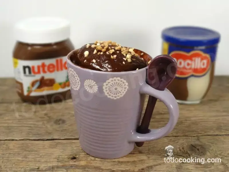 Taza lila con mug cake de chocolate sobre mesa de madera rústica, con botes de Nutella y Nocilla al fondo para mostrar los ingredientes.