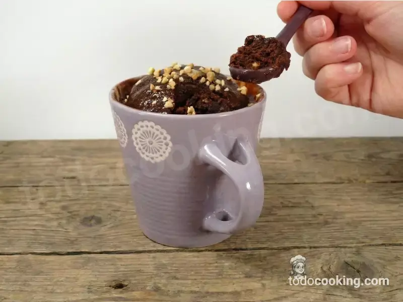 Primer plano de una mano sosteniendo una cucharada de mug cake de chocolate esponjoso con trozos de almendra, mostrando la textura interior del bizcocho.