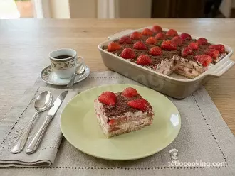 Porción de tiramisú de fresas servida en un plato junto a una taza de café, con la bandeja completa al fondo.
