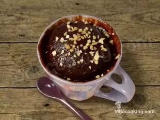 Vista cenital de un mug cake de chocolate recién hecho en microondas, cubierto con topping de almendras picadas dentro de una taza lila.